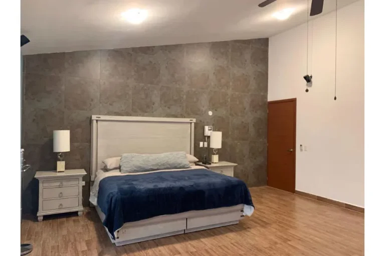 Recámara con cama de cabecera clara, cobertor azul, burós con lámparas y muro de piedra detrás de la cama.