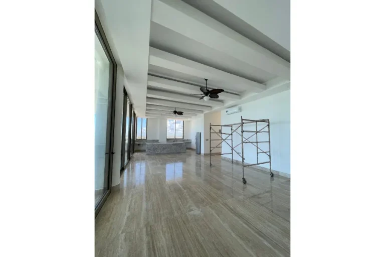 Sala sin muebles con grandes ventanales, piso de piedra pulida, vigas en el techo, ventiladores y un andamio.