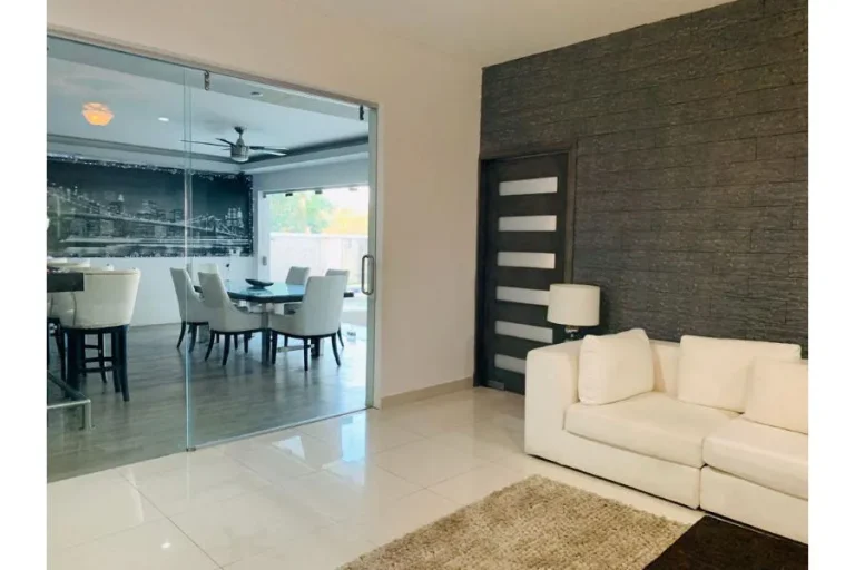 Sala con sofá blanco, alfombra y muro oscuro texturizado, junto a un comedor separado por puertas de cristal.