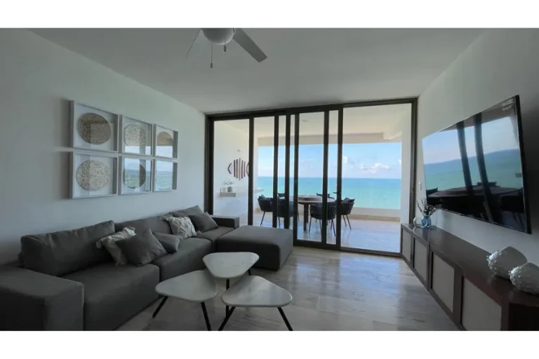 Sala con sofá gris, mesas de centro y televisión, conectada por puertas corredizas a una terraza con vista al mar.