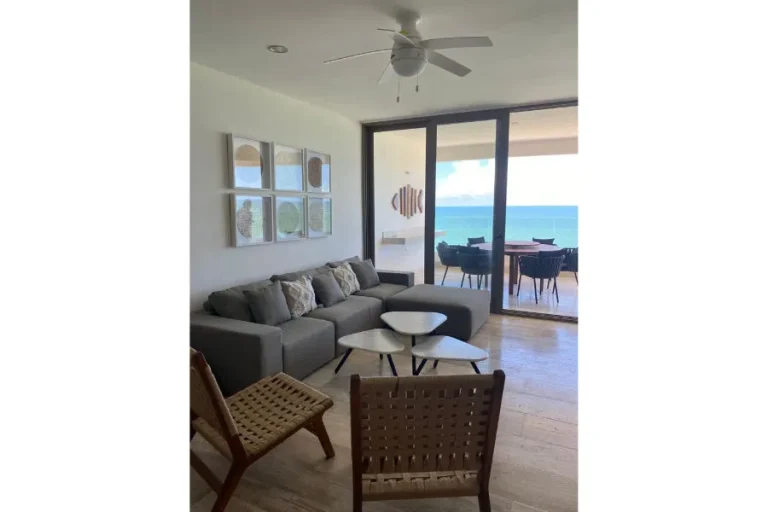 Sala con sofá gris, sillas tejidas y mesas pequeñas, junto a puertas de cristal hacia una terraza con vista al mar.