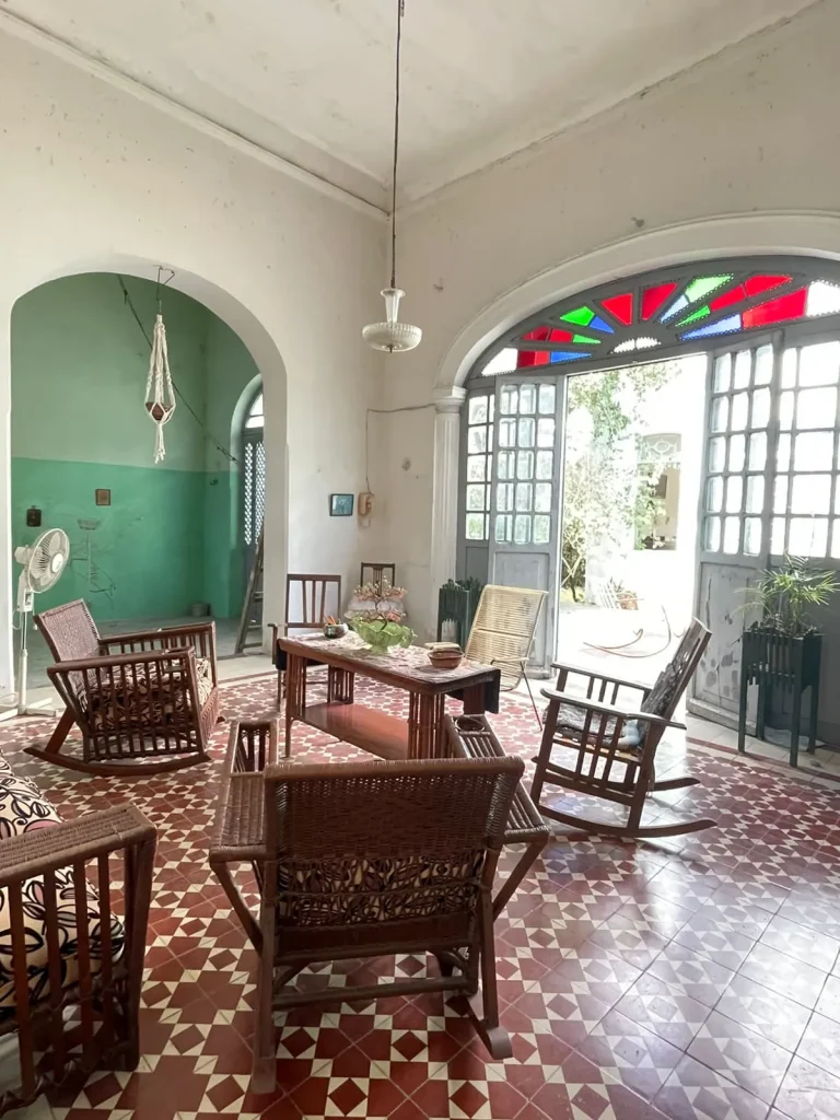 Sala con mecedoras de madera, piso geométrico y una puerta de arco abierta con vidrios rojos y verdes en la parte superior.