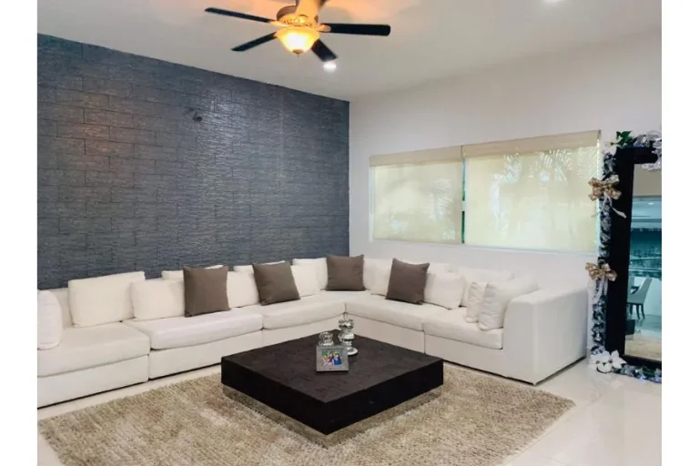 Sala con sofá blanco en L, cojines oscuros, mesa de centro cuadrada, alfombra y muro gris con textura.
