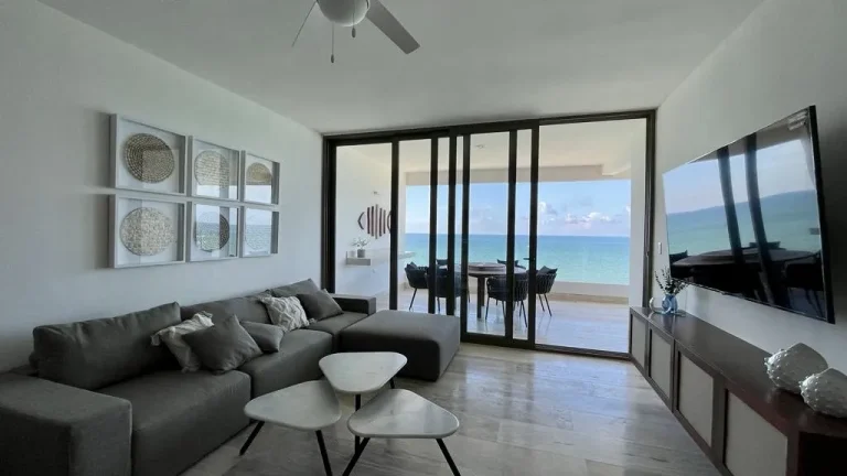 Sala con sofá gris y televisor, conectada por puertas corredizas de cristal a una terraza con vista al mar.