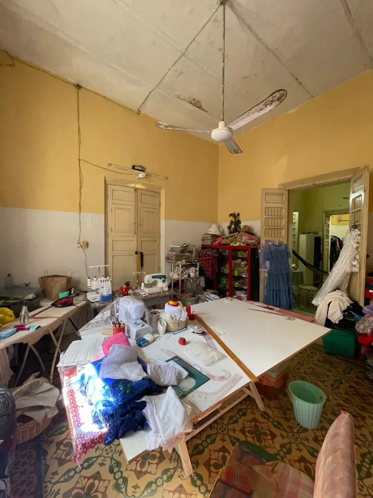 Taller de costura con mesa de corte, máquinas de coser, telas y vestidos, paredes amarillas y ventilador de techo.