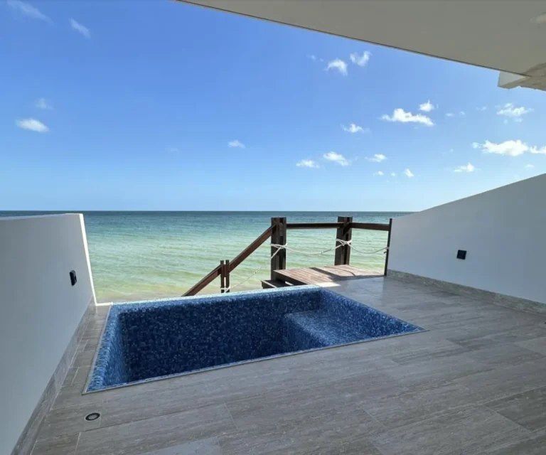 Terraza con alberca revestida de mosaico azul, piso de loseta y escalera de madera hacia el mar.