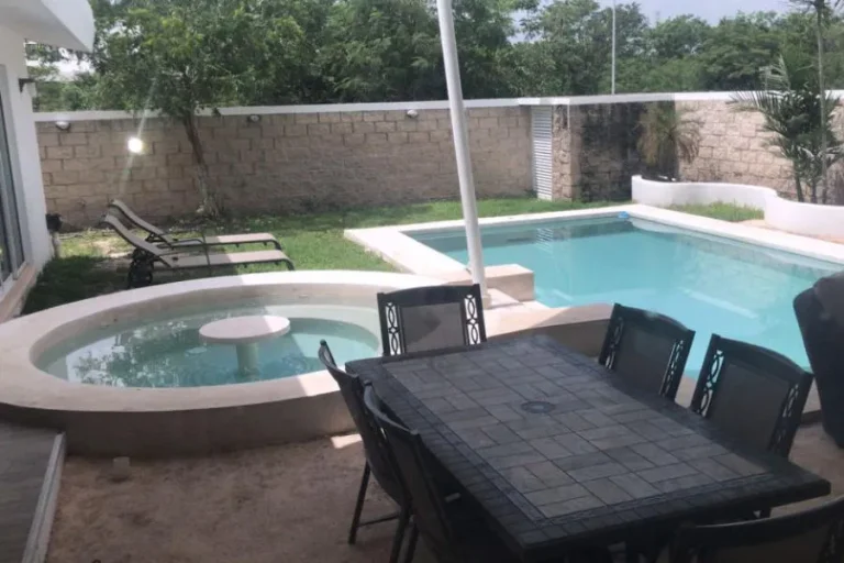 Terraza con mesa y sillas, alberca rectangular, piscina circular contigua y camastros junto al jardín.