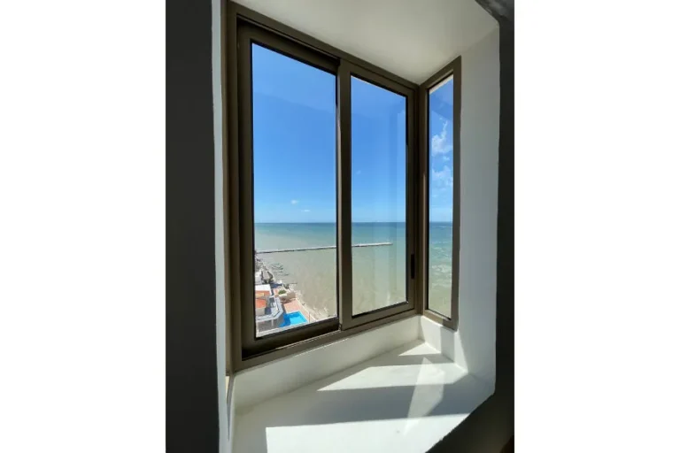 Ventana de marco oscuro con una hoja abierta, vista al mar y luz solar sobre el alféizar blanco.