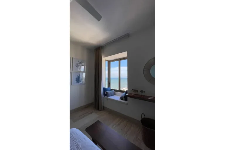 Rincón con asiento bajo una ventana con vista al mar, cojines azules, cortina beige y espejo redondo en la pared.