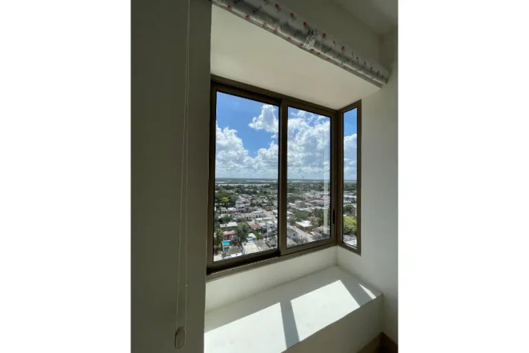 Ventana de esquina con vista a edificios y vegetación, cielo con nubes y luz solar sobre el antepecho.