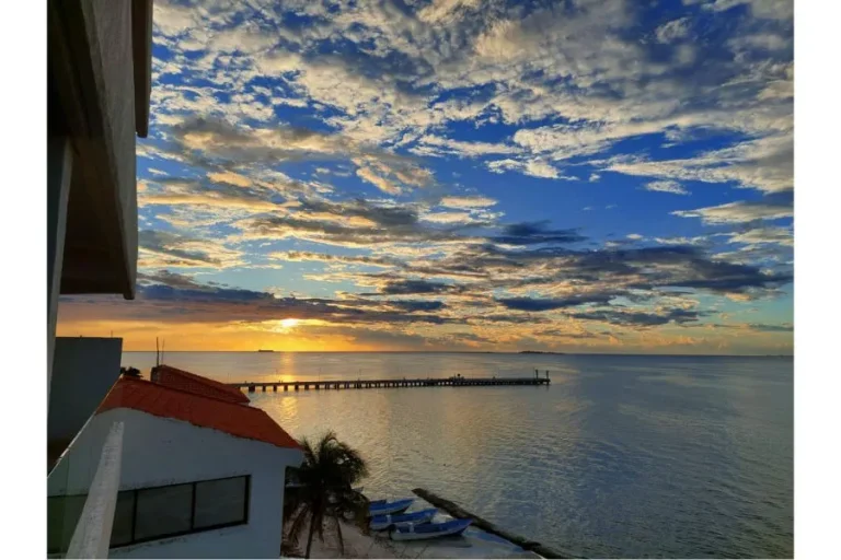 Sol cerca del horizonte sobre el mar, con nubes iluminadas, un muelle y una construcción en primer plano.